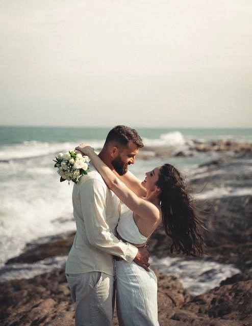 Casal abraçado à beira-mar segurando um buquê, no fim da tarde.